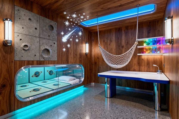 A kids’ bedroom with walnut wood walls and ceiling features a clear acrylic pod bed with teal cushions printed with planets, lit by cyan LED strip lighting. A cast-concrete wall panel resembles the moon’s craters, with star and comet decals nearby. A white desk with blue edging and chrome legs sits beside floating shelves displaying rainbow acrylic blocks. A rope hammock hangs from the ceiling. Polished gray terrazzo floors and tubular chrome wall sconces complete the space.
