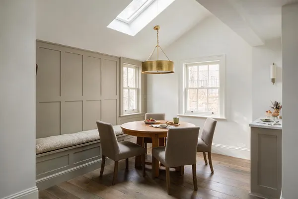 A bright transitional sunroom with a vaulted white ceiling, skylight, and brass drum chandelier above a round wood table surrounded by four upholstered taupe chairs. The room features a built-in bench with a cushioned seat, paneled taupe wall treatment, white-trimmed windows, and warm wide-plank wood flooring.