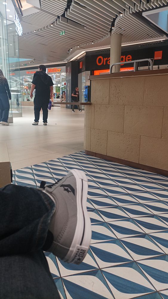 A man wearing a mask sits on a bench in a shopping area, using his smartphone while waiting.
