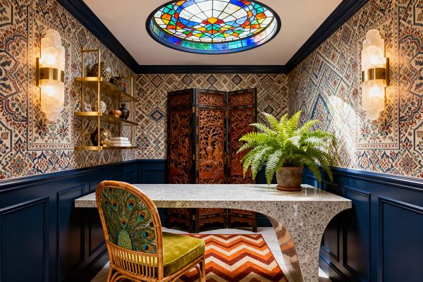 A jewel-toned maximalist home office with navy paneled wainscoting and patterned wallpaper. A circular stained-glass skylight illuminates a curved terrazzo desk topped with a potted fern. Two alabaster-and-brass sconces flank floating brass shelves styled with ceramics and books. At the back, an intricately carved wooden folding screen stands behind a rattan chair with a green seat and peacock-patterned back, over a rust chevron rug.