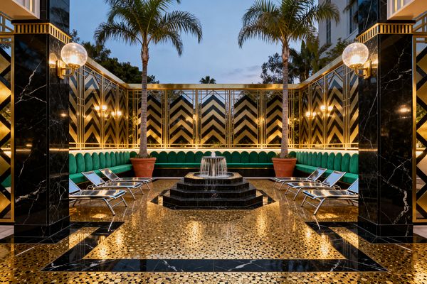 A symmetrical Art Deco patio features a central tiered black marble fountain on a gold mosaic floor bordered by black stone. Brass-framed chevron screens and globe sconces line the walls above a continuous emerald velvet banquette, with chrome sling loungers on both sides. Two potted palm trees rise behind the seating, and dusk lighting casts a warm glow across the reflective gold and black surfaces.