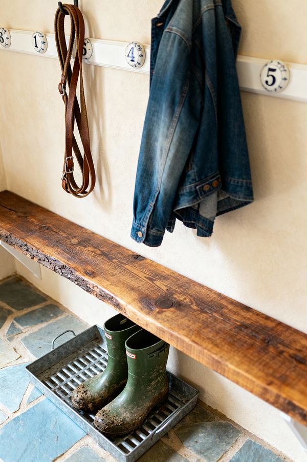 A farmhouse mudroom vignette shows a wall-mounted live-edge wooden bench against warm cream plaster walls. Above, a white peg rail with black hooks and blue-and-white numbered ceramic disks holds a denim jacket and a looped leather strap; below, a galvanized boot tray cradles muddy green rubber boots on a blue-gray flagstone floor, emphasizing durable materials and a simple, organized layout.