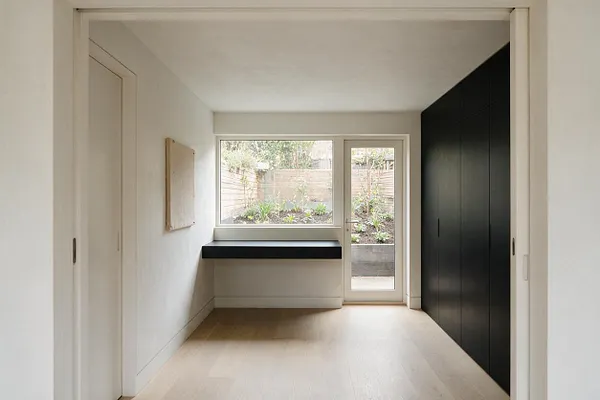 A minimalist backyard room with white walls, light wood flooring, and a large window and glass door opening to a small landscaped garden. A slim black built-in desk or shelf sits beneath the window, and a full-height matte black cabinet wall lines the right side.