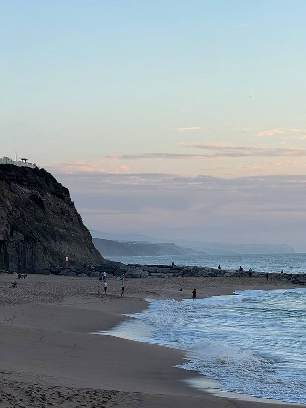 A serene beach scene at dusk with people walking along the shore and rocky formations in the background.