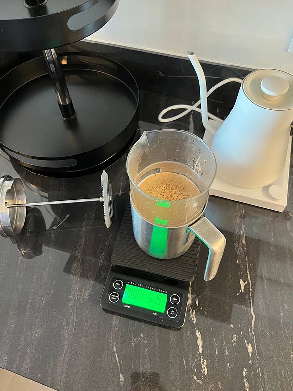 A coffee preparation setup featuring a French press, a kettle, and a digital scale on a marble countertop.