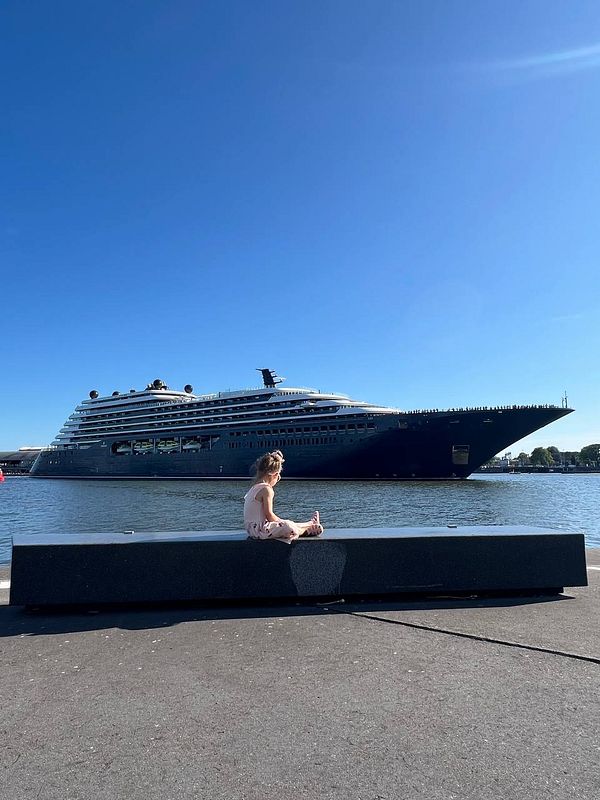 A child sits on a stone bench by the water, with a large yacht in the background under a clear blue sky.
