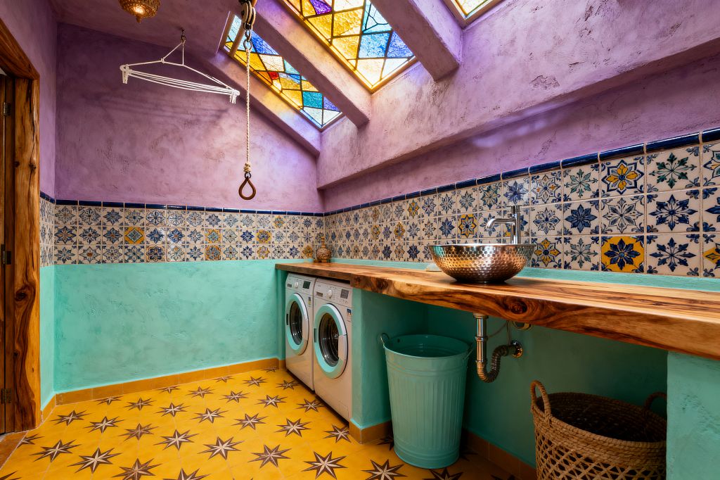 A bohemian laundry room features lavender plaster walls above aqua stucco and a band of blue-and-yellow patterned Talavera tiles, lit by sloped skylights with jewel-toned stained glass. A live-edge wood counter spans a front-load washer and dryer and holds a hammered metal vessel sink with a chrome faucet, while a bright yellow star-pattern floor, woven basket, mint metal bin, a white ceiling drying rack, and a rope-and-pulley detail add functional character.