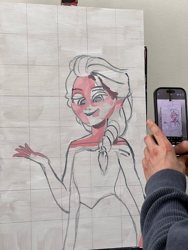A partially completed acrylic painting of a character with a light background and a person holding a phone to capture the work.