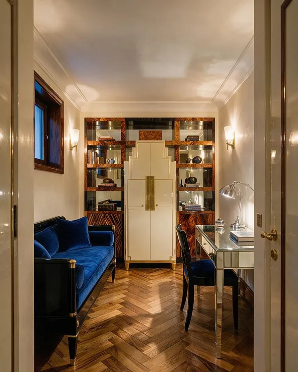 A narrow guest bedroom styled in Art Deco fashion features a deep blue velvet sofa on the left, a mirrored writing desk with a chrome lamp on the right, and a cream cabinet centered against the back wall. The room has warm herringbone wood flooring, cream walls, and built-in shelving with glossy wood and mirrored panels, creating a refined and symmetrical look.