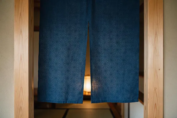 A Japanese Zen-style doorway framed in pale natural wood with textured blue noren curtains hanging in the center. A warm lantern glow is visible behind the fabric, with a calm tatami-lined interior beyond.