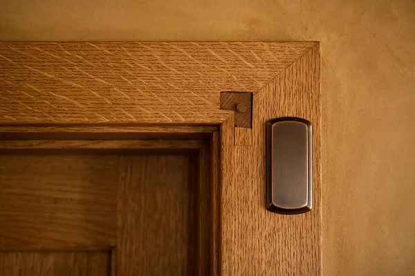 Close-up of a Craftsman-style wooden door frame with prominent corner joinery and a textured oak grain. A slim brushed metal sensor or doorbell is mounted on the wall beside the trim against a warm beige plaster background.