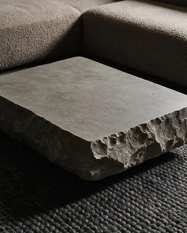 A low rectangular stone coffee table with a smooth top and rough, chiseled sides sits in front of a textured dark sofa and woven rug. The muted gray surface and rugged edges create a heavy, sculptural Brutalist look.