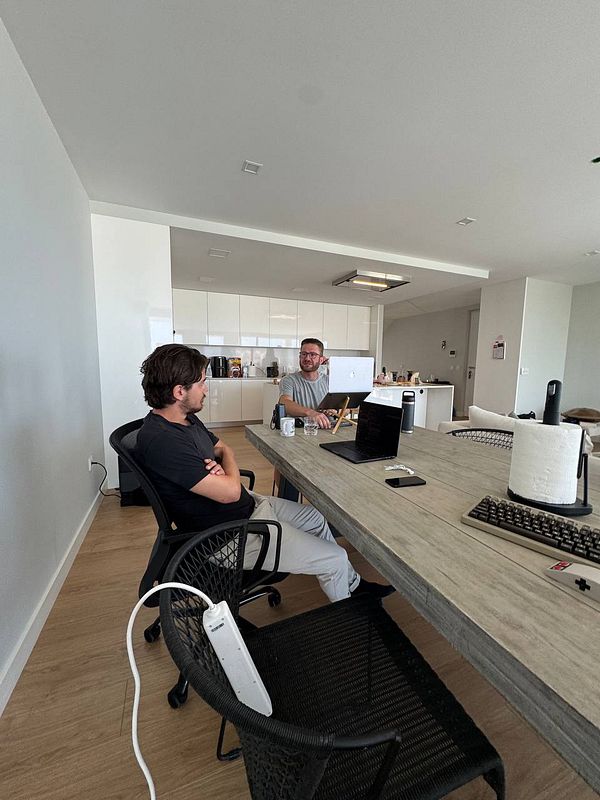 Two men are seated at a large table in a modern coworking space, engaged in conversation.