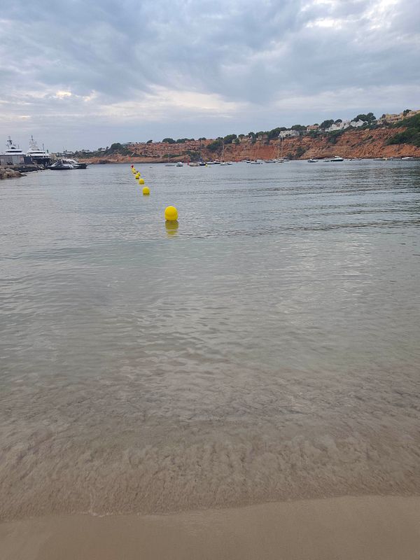 Calm sea shore with yellow buoys marking a swimming area, boats anchored near a rocky cliff, and a cloudy sky.