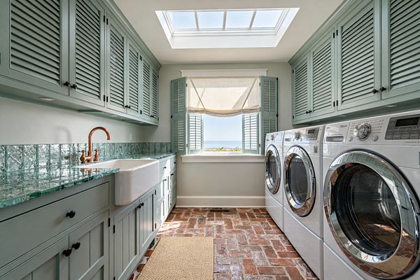 A narrow coastal laundry room with seafoam green louvered cabinets above and below, a thick aqua glass countertop and matching textured glass backsplash, and a white farmhouse sink with a copper faucet. Four front-loading machines line the opposite wall, while a large skylight and a window with matching shutters and a soft roman shade brighten the tumbled brick floor and natural fiber rug, with the ocean visible beyond.