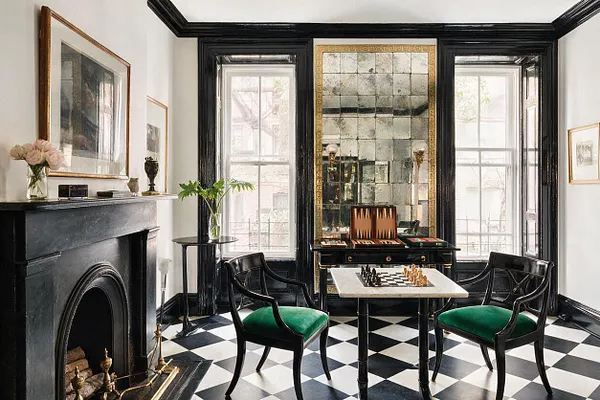 A Hollywood Regency game room with black lacquer trim, tall mirrored wall panels, and a black-and-white checkered floor. A small game table with chess pieces sits between two curved black chairs with green velvet seats, with large windows and a dark fireplace adding symmetry and drama.