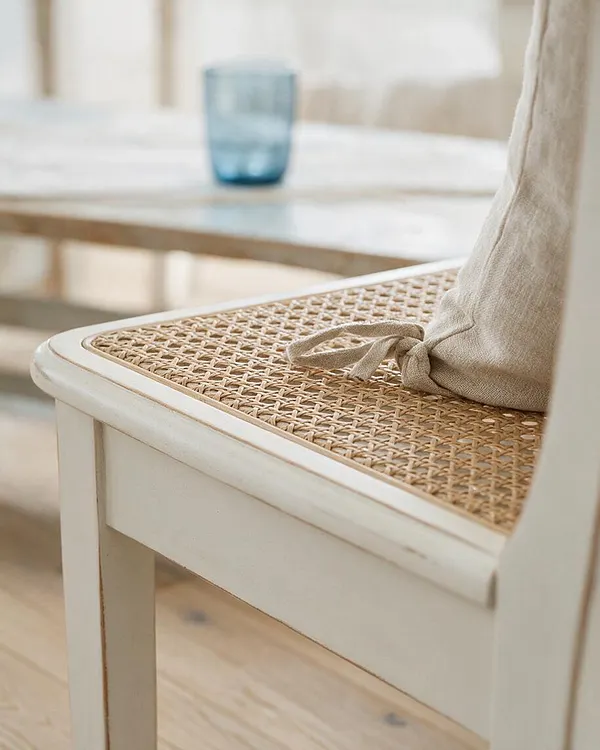 A close-up of a painted dining chair with a woven cane seat and a tied natural linen cushion. In the blurred background, a light wood table holds a translucent blue glass, reinforcing the airy coastal look.