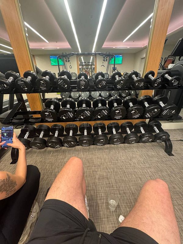 A gym scene featuring a rack of dumbbells and a person seated nearby.