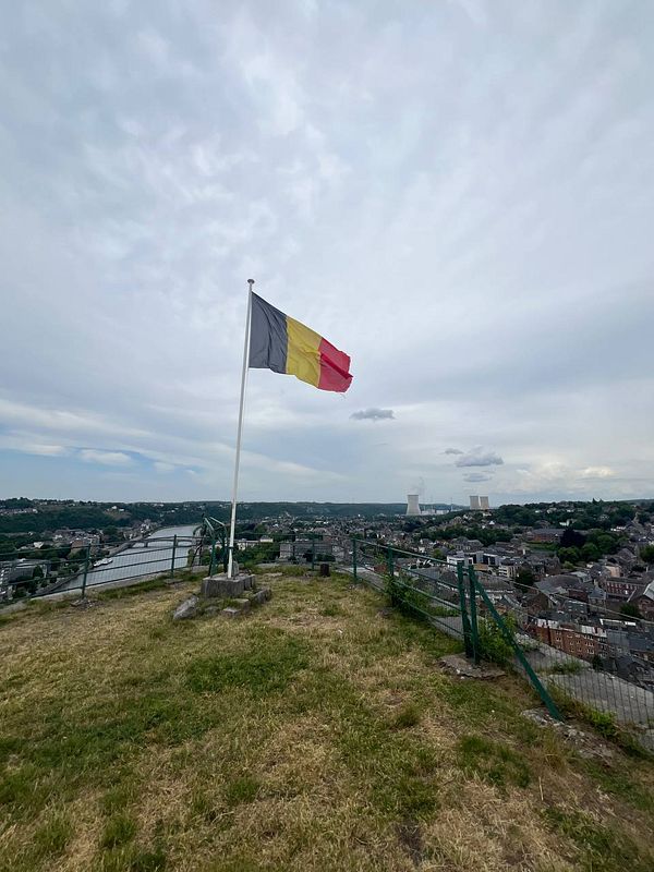A Belgian flag waves atop a hill overlooking a scenic view of a town and river.