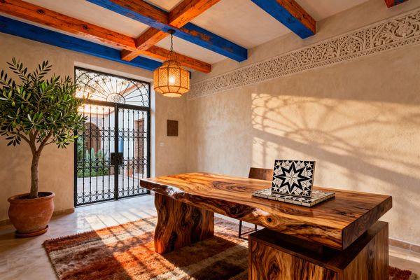 A Moroccan-inspired home office with sand-colored plaster walls and a carved frieze features vivid blue and burnt-orange ceiling beams and a perforated brass pendant casting patterned light. A large live-edge wood desk sits on a rust and cream shag rug near wrought-iron double doors opening to a sunlit courtyard; a terracotta pot with an olive tree stands to the left, and a black-and-white zellige star tile rests on the desk.