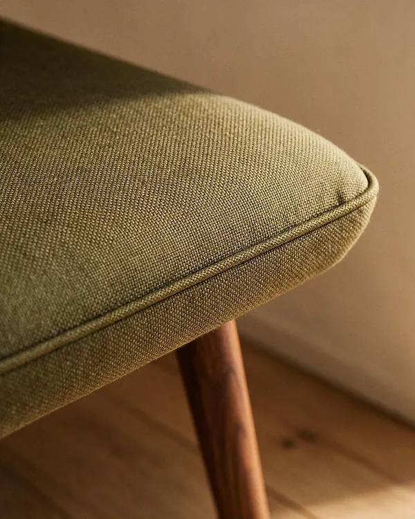 Close-up of a mid-century modern upholstered seat in olive green fabric with fine woven texture and tailored piping along the edge. A tapered walnut leg is visible beneath the cushion, with warm wood flooring and a neutral wall in the background.