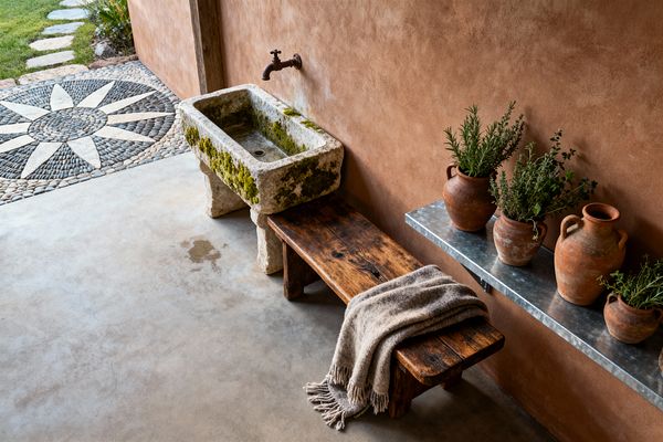 An outdoor patio with a warm terracotta plaster wall features a mossy, weathered stone trough sink beneath a rusty wall-mounted spigot, a solid reclaimed wood bench topped with a fringed wool throw, and a galvanized metal shelf holding terracotta pots of herbs. Smooth concrete flooring opens to a garden path where a circular pebble mosaic forms a star pattern and stepping stones lead across a lawn, all lit by natural light that highlights the earthy textures and tones.
