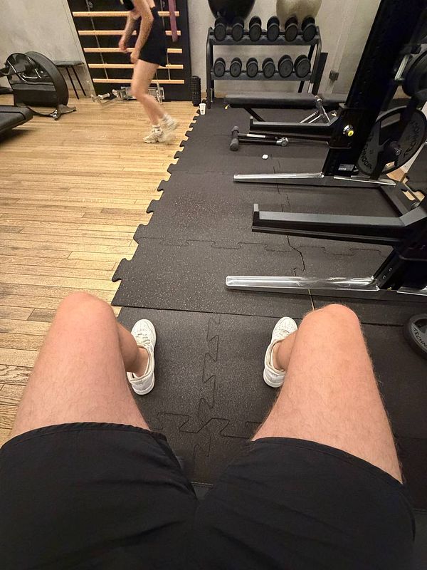 View from seated person at gym showing legs and workout area with equipment and a woman walking in the background.