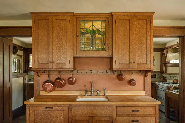 A Craftsman-style kitchen featuring warm oak cabinets, a central sink area, and a stained glass cabinet panel above the backsplash. Copper pots hang from black hooks on a rail, complementing the earthy plaster wall and wood-trimmed doorways. The room has a classic, handcrafted feel with natural finishes and soft, ambient light.