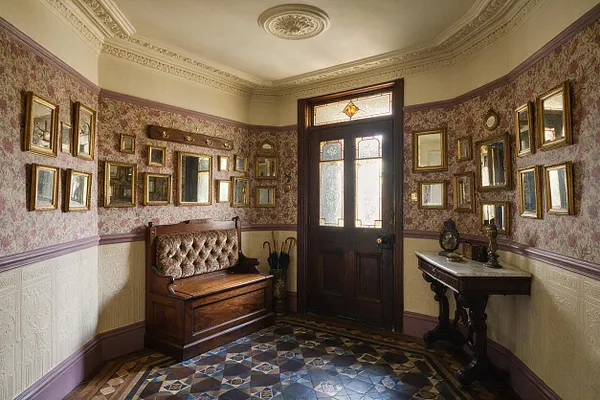 A Victorian-style front porch or entry vestibule with floral wallpaper, cream wainscoting, and a dark wood door with frosted glass and stained-glass detail. The space is filled with many gold-framed mirrors and pictures, a tufted wooden bench, a small pedestal table, and a patterned tile floor.