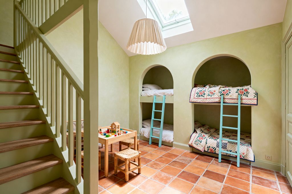 A children's room in French Country style with sage green limewashed walls features two built-in arched alcoves housing stacked bunks. Patchwork floral quilts and white bedding sit behind pale turquoise ladders. Rustic terracotta floor tiles, a natural wood play table with woven rush-seat stools, a green-painted staircase, a skylight, and a pleated fabric pendant complete the warm, sunlit space.