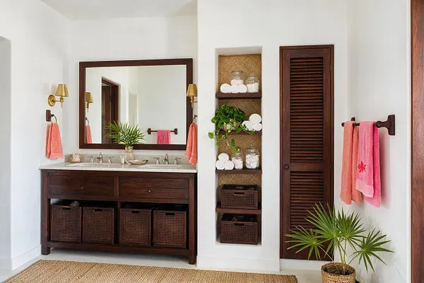 A tropical-style master bathroom with a dark wood vanity, large framed mirror, and brass wall sconces flanking the sink. Coral towels add color against crisp white walls, while woven storage baskets, a louvered wood door, and potted palms bring natural texture and a resort-like feel.