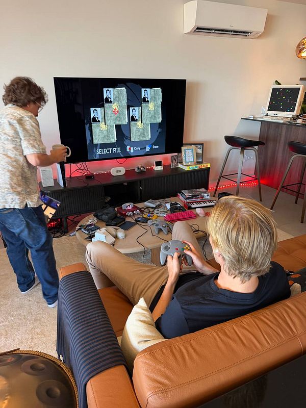 Two young men playing and setting up video games in a modern living room with retro gaming consoles and a large TV display.