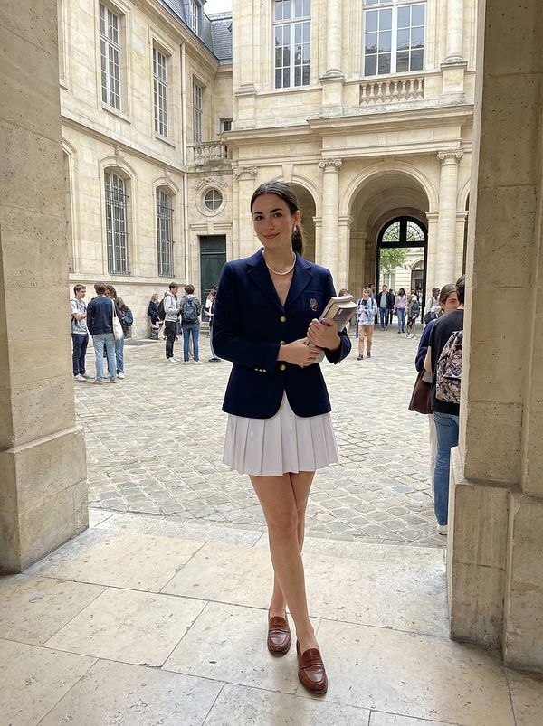 A young woman stands confidently in a courtyard, dressed in a stylish outfit, surrounded by people.