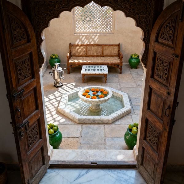 View through ornate carved wooden double doors into a small traditional courtyard patio. An octagonal white-marble fountain with floating orange marigolds centers pale stone pavers. A wooden bench with cane seating lines the back wall beneath a decorative lattice screen, with a low green-and-white marble-inlay table in front. Green glazed urns filled with citrus flank the space, and a polished metal samovar sits to the left. Sunlight filters through the lattice, casting patterned shadows.