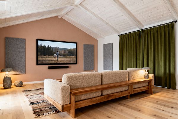 A Scandinavian media room features a terracotta plaster wall with a wall-mounted TV and slim black soundbar, flanked by gray felt acoustic panels. A solid oak sofa with beige bouclé cushions sits on light oak floors beside a striped flatweave rug; olive blackout curtains cover the window wall, a whitewashed vaulted wood ceiling spans overhead, and two stone lamps provide warm, soft accent lighting.