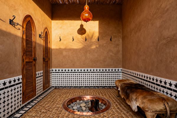 Moroccan-style mudroom with warm terracotta plaster walls above a wainscot of white zellige tiles patterned with black stars, two arched louvered wooden doors, and a row of black wrought-iron hooks over a straw-colored woven floor edged in black-and-white tile. At the center is a recessed round copper basin filled with river stones and boots, lit by an amber glass pendant from a slatted reed ceiling, with a cowhide-draped bench on the right.