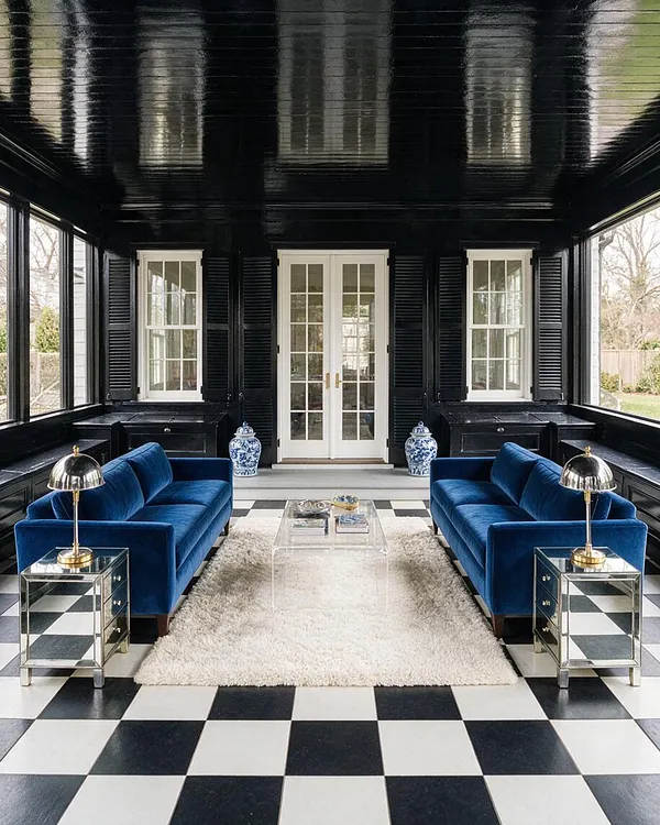 A dramatic front porch enclosed in glossy black paneling and ceiling, with French doors centered between white-trimmed windows and shutters. Two cobalt blue velvet sofas face each other over a white shag rug and clear acrylic coffee table, while mirrored side tables and blue-and-white porcelain jars add Hollywood Regency glamour.