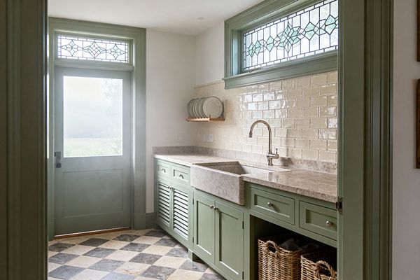 A traditional laundry room features sage green shaker cabinets with a louvered door, a thick beige stone apron-front sink with matching countertop, and a glossy cream subway tile backsplash. Light filters through a leaded stained-glass transom and a matching green door, while a charcoal-and-ivory checkerboard stone floor, polished nickel knobs, a small wooden plate rack, and woven baskets complete the bright, orderly workspace.