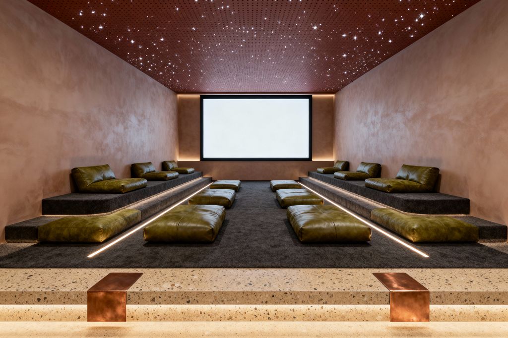 A modern media room with tiered platforms covered in dark charcoal carpet features olive green leather lounge cushions along both sides and low floor pads down the center. Terracotta plaster walls frame a large black-trimmed screen with subtle cove lighting, while a rust-toned perforated ceiling dotted with star-like fiber-optic lights adds ambience. Terrazzo entry steps with copper details and integrated LED step lights complete the warm, cinematic setting.
