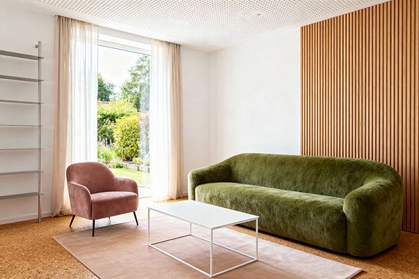 A modern living room with a long moss-green plush sofa set before a vertical oak slat wall. A dusty rose velvet armchair sits near a floor-to-ceiling window with sheer curtains, facing a slim white metal coffee table on a blush rug over cork flooring. A tall metal shelving unit stands at the left, and a perforated white ceiling tops the bright space that looks out to a garden.
