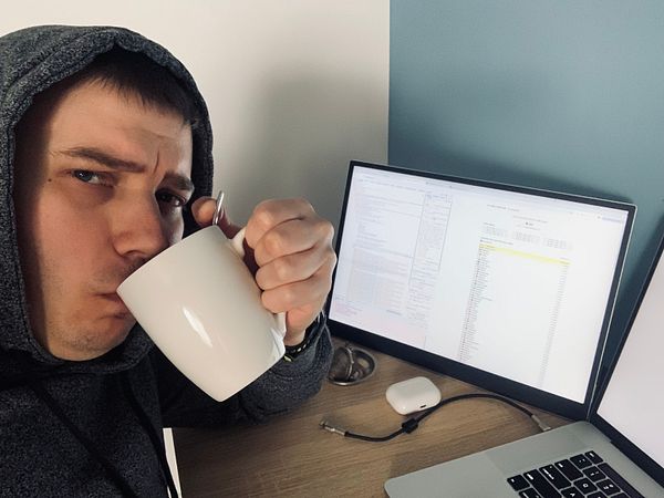 A person in a hoodie is sipping from a white mug while working at a desk with multiple screens.