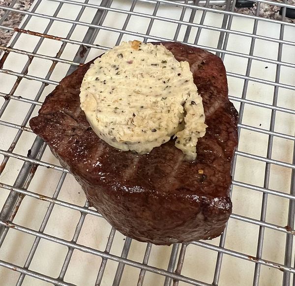 A cooked steak topped with a round portion of herb butter on a wire rack.