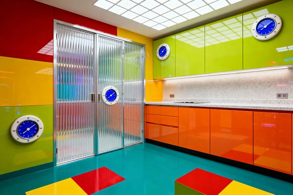 A laundry room with bold, glossy color blocking shows lime green upper cabinets and orange lower drawers under a speckled white countertop with continuous under-cabinet lighting. A teal high-gloss floor with red and yellow squares meets ribbed fluted-glass sliding doors in aluminum frames, while round blue clocks punctuate the walls and cabinetry, and a luminous grid ceiling evenly lights the space.