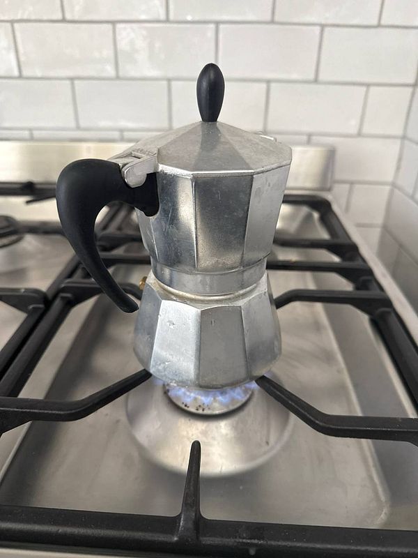 A stovetop espresso maker is placed on a gas burner, with flames visible underneath.