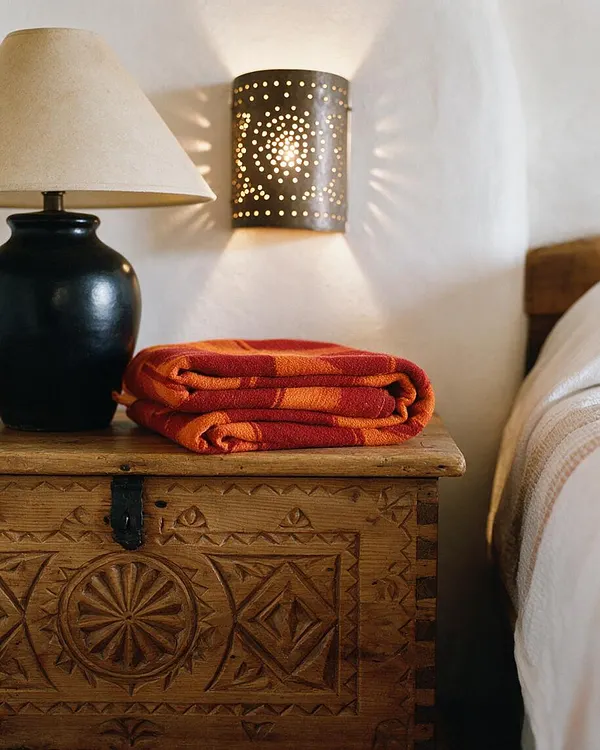 A Southwestern-style bedroom vignette shows a carved wooden chest used as a nightstand beside a bed with white bedding. On top sit a black ceramic lamp with a beige shade and a folded red-orange textile, while a pierced metal wall sconce casts patterned light on a white wall.