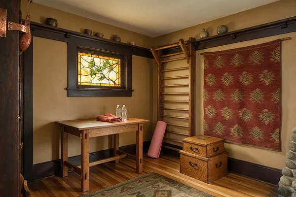 A compact Craftsman-style home gym with tan walls, dark wood trim, and a stained glass window above a simple wooden table. Wall-mounted ladder bars, a rolled yoga mat, stacked wooden storage boxes, and woven textile wall art add warmth and character to the workout space.