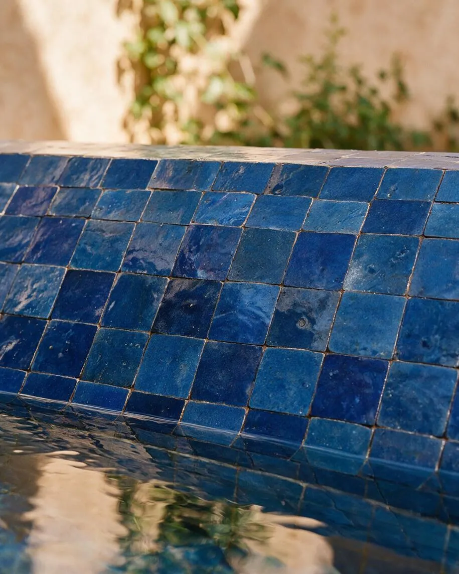 A close-up of a curved pool edge covered in small square blue Moroccan-style tiles with varied glossy finishes. The water below reflects the tiles and surrounding greenery against a warm, blurred background.