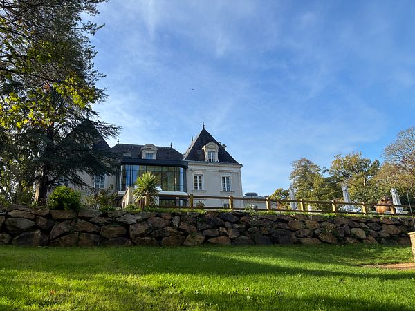 Elegant castle-like hotel with a white facade, steep dark roofs, and a glass extension, surrounded by a lush garden and stone wall under a bright blue sky.