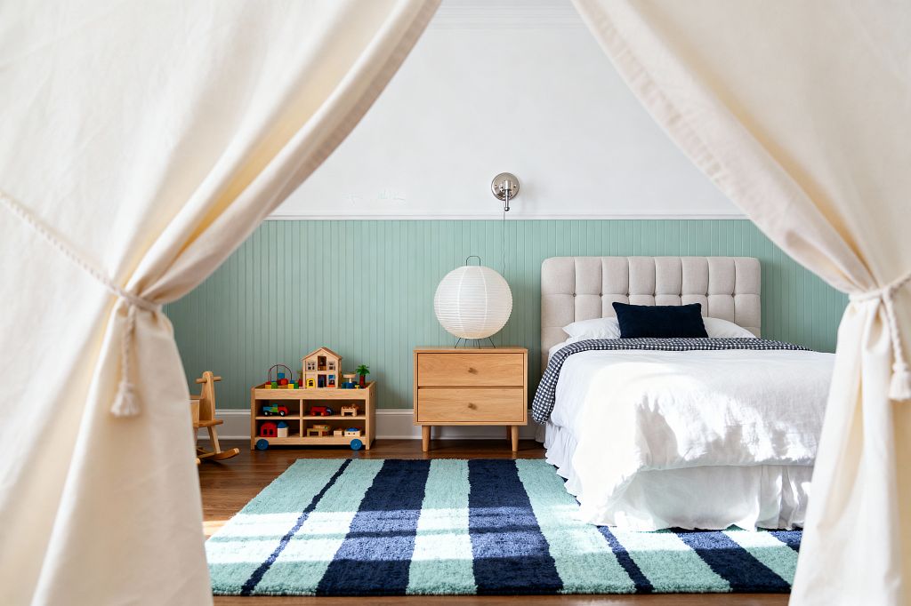 View through the opening of a cream canvas play tent into a children’s bedroom with seafoam beadboard wainscoting and white upper walls. A light beige tufted headboard and white bedding sit on a striped aqua-and-navy rug, accented by a navy pillow and gingham blanket. A light oak nightstand holds a round paper lantern beneath a brushed-nickel wall sconce, with a low wood toy shelf and warm hardwood floors completing the scene.