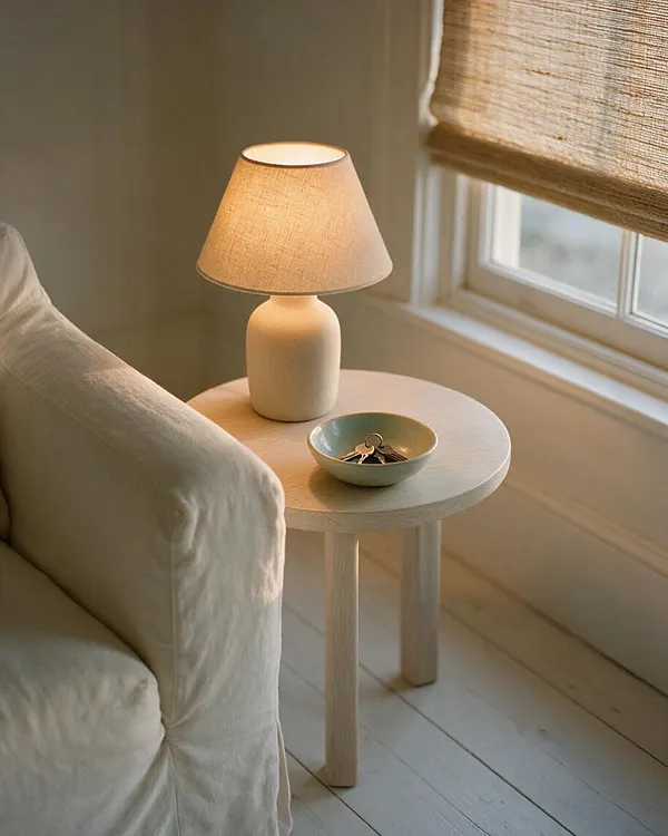 A cozy coastal-style reading nook with a slipcovered armchair beside a small round light wood side table. The table holds a ceramic bowl with keys and a small ceramic lamp with a linen shade, set next to a window with a woven Roman shade that filters warm daylight.